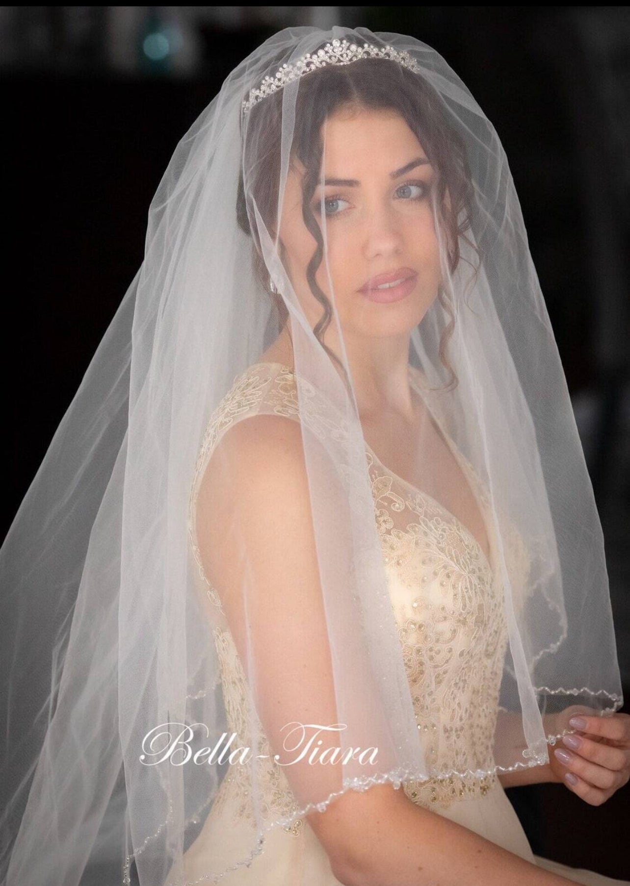A bride wearing a white cathedral veil with beaded pearl and crystal trim, and a blusher layer, in front of a dark background.
